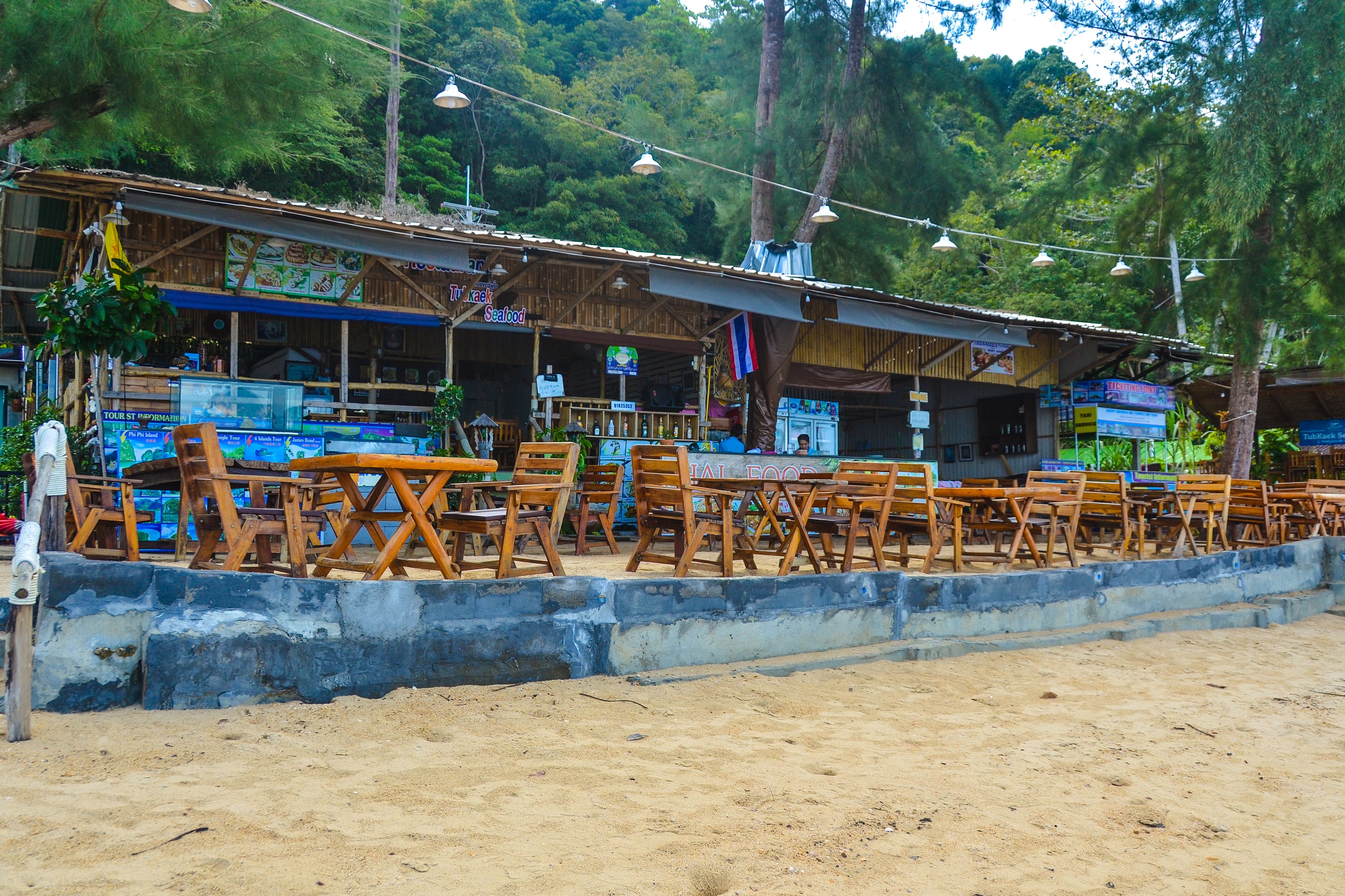A beachside restaurant with outdoor seating, wooden tables and chairs, and a sign indicating seafood cuisine.