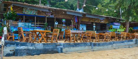 A beachside restaurant with outdoor seating, wooden tables and chairs, and a sign indicating seafood cuisine.