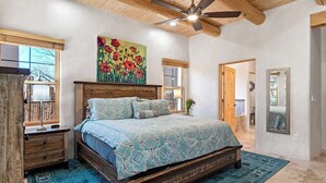 Une chambre avec un lit en bois, un ventilateur de plafond et une grande fenêtre donnant sur les arbres.