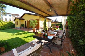 Un patio al aire libre con una mesa dispuesta para comer, rodeado de plantas en macetas y un jardín.