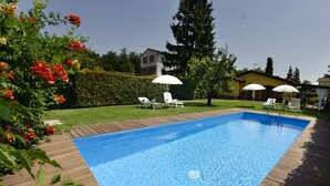 Une piscine rectangulaire entourée d’une terrasse en bois. On aperçoit, au fond, des chaises longues blanches et des parasols.