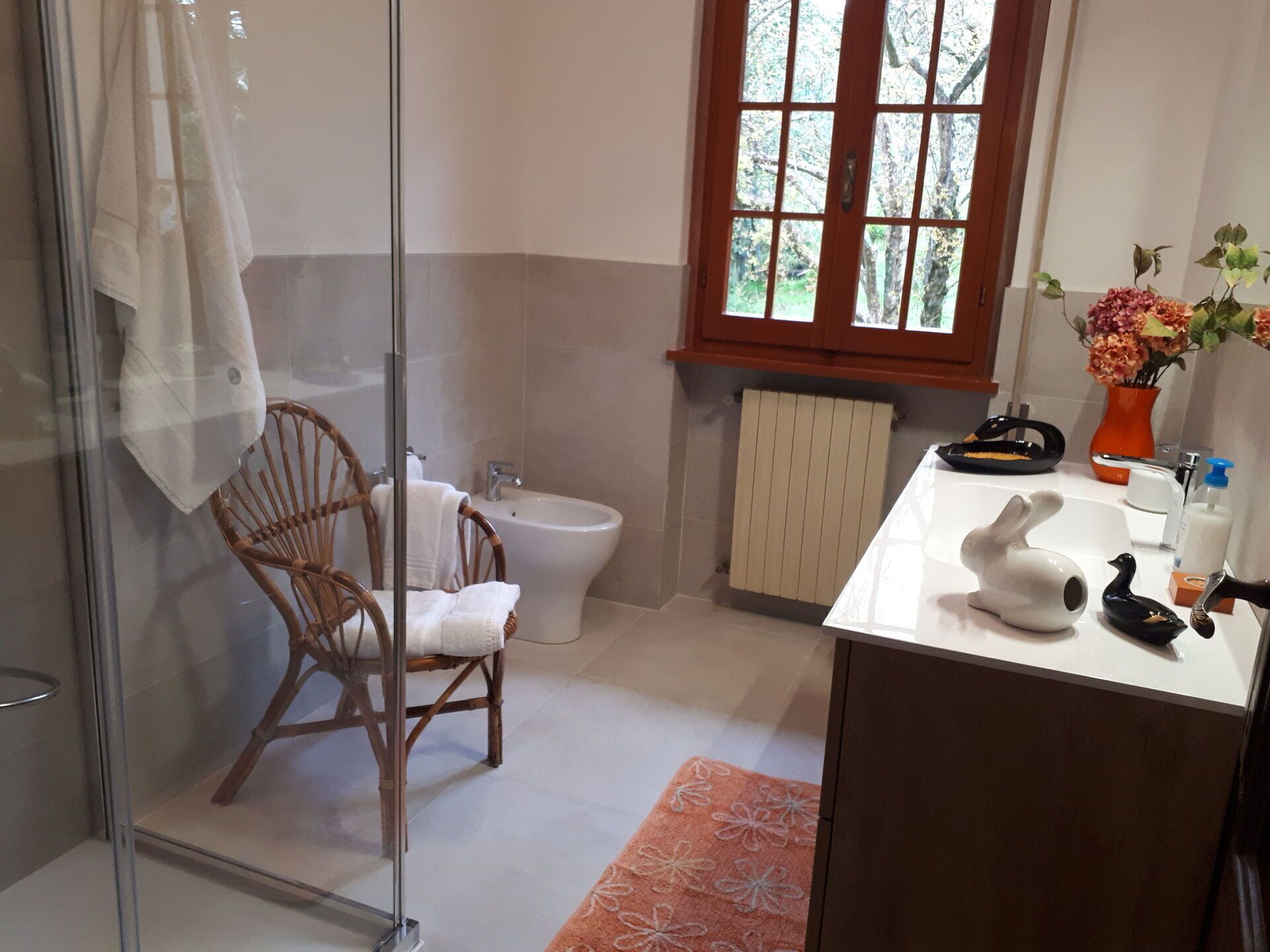 A bathroom with a glass shower enclosure, a white toilet, a wooden chair, a white sink with a duck-shaped soap dispenser, and a window with a view of trees.