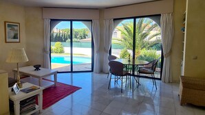A room with a dining table and chairs, a small white table with a lamp, a red rug, and a view of a swimming pool and greenery outside. - Dunas Douradas Beach Villa (Vale do Lobo)