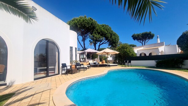A swimming pool with lounge chairs and umbrellas, surrounded by white buildings and trees. - Dunas Douradas Beach Villa (Vale do Lobo)