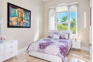 Un dormitorio con cama, mesita de noche, ventana con vista a palmeras y un cuadro en la pared.