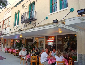 An outdoor seating area with tables and chairs, a balcony with a railing, and a building facade with windows and a neon sign.