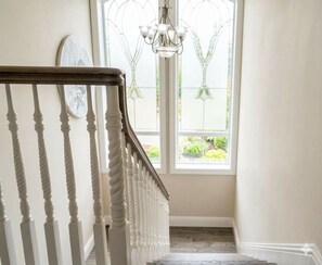 A staircase with white balusters leading to a landing, a chandelier above, and a window with a view of greenery.
