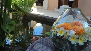A glass vase with flowers on a round table by a water feature, with a building and a person in the background. - Villa Pasraman Baghawan (Jimbaran)