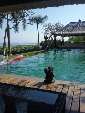 A pool with a view of the ocean, surrounded by palm trees and a thatched roof structure. - Villa Pasraman Baghawan (Jimbaran)