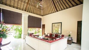 A spacious living area with a white sofa, red cushions, and a large window with a view of greenery. - Villa Pasraman Baghawan (Jimbaran)