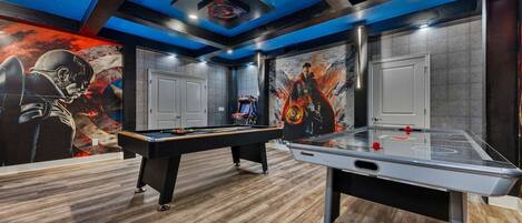 Una mesa de billar junto a una mesa de tenis de mesa, un techo con iluminación azul y una pared con un mural.