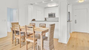 Un coin repas avec une table en bois et des chaises en osier, des murs blancs et une cuisine dotée de meubles blancs et d’un four à micro-ondes.