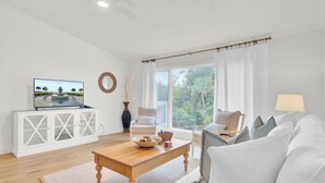 Un salon spacieux avec un canapé blanc, une table basse en bois et un téléviseur posé sur un meuble blanc. Il y a une fenêtre avec des rideaux, une vue sur la verdure à l’extérieur et une lampe sur une table d’appoint.
