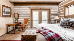 Un dormitorio de cabaña de madera con cama, sillón, lámpara y ventana.