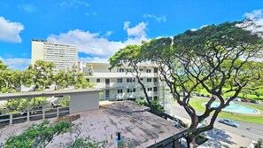 A multi-story building with balconies, a parking area, and a large tree in the foreground.