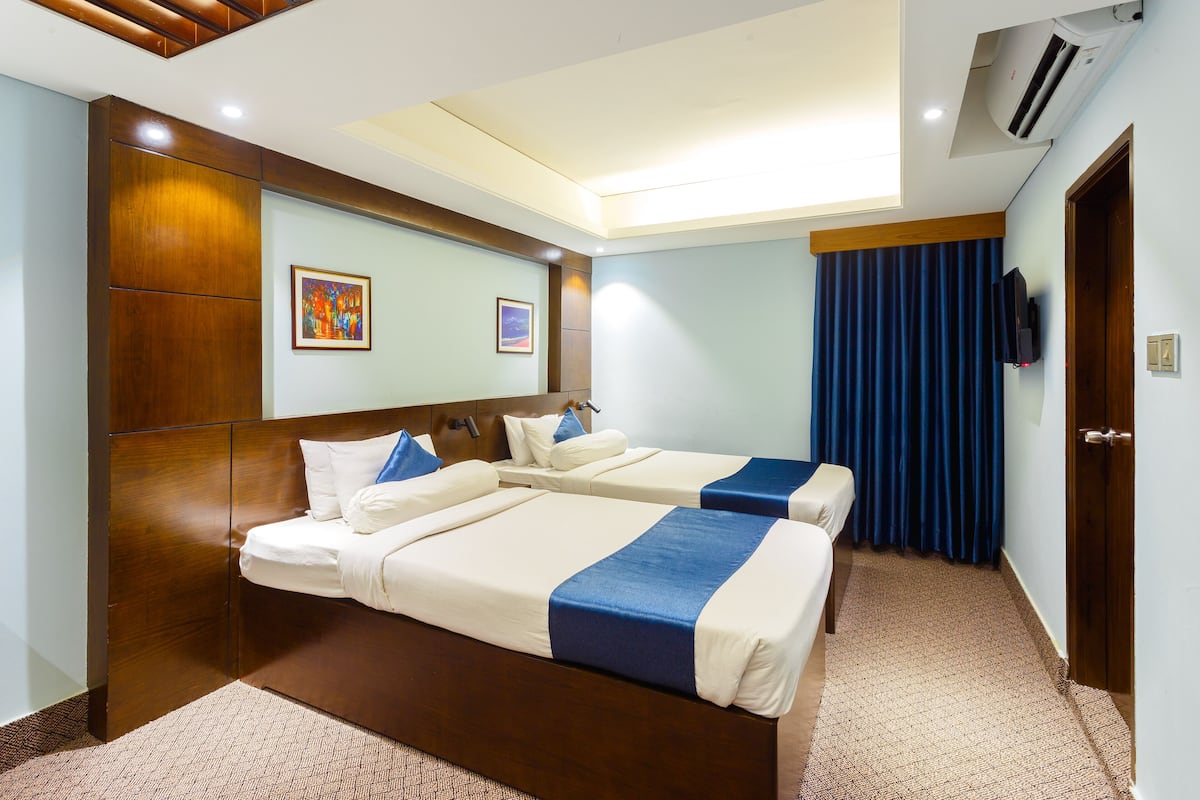 A hotel room with two beds, a wooden headboard, a ceiling light, a painting, a TV, and a blue curtain.