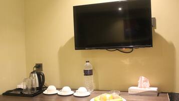 Ein Hotelzimmer mit Flachbildfernseher an der Wand, einer Kaffeemaschine, Tassen, einer Wasserflasche, einem Teller mit Obst und einer Taschentuchbox.