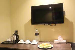 A hotel room with a flat-screen TV mounted on the wall, a coffee maker, cups, a bottle of water, a plate with fruit, and a tissue box. - Hotel The Capital (Dhaka)