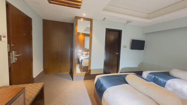 A hotel room with a wooden wardrobe, a bed with blue bedding, a bedside table, a television mounted on the wall, and a carpeted floor. - Hotel The Capital (Dhaka)