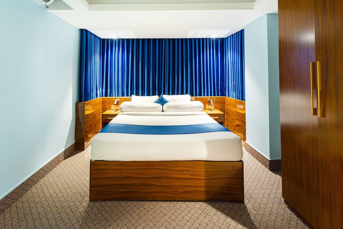 A hotel room with a large bed, wooden headboard, and a blue curtain backdrop.