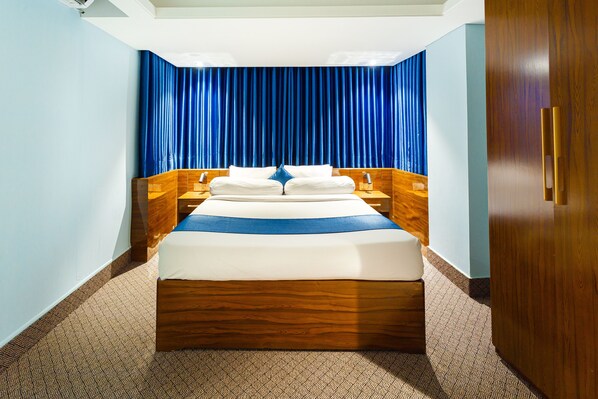 A hotel room with a large bed, wooden headboard, and a blue curtain backdrop. - Hotel The Capital (Dhaka)