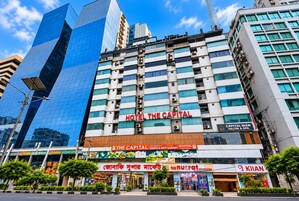 Un hotel de varios pisos con un cartel llamativo que dice 'HOTEL THE CAPITAL' y diversos establecimientos comerciales en la planta baja.