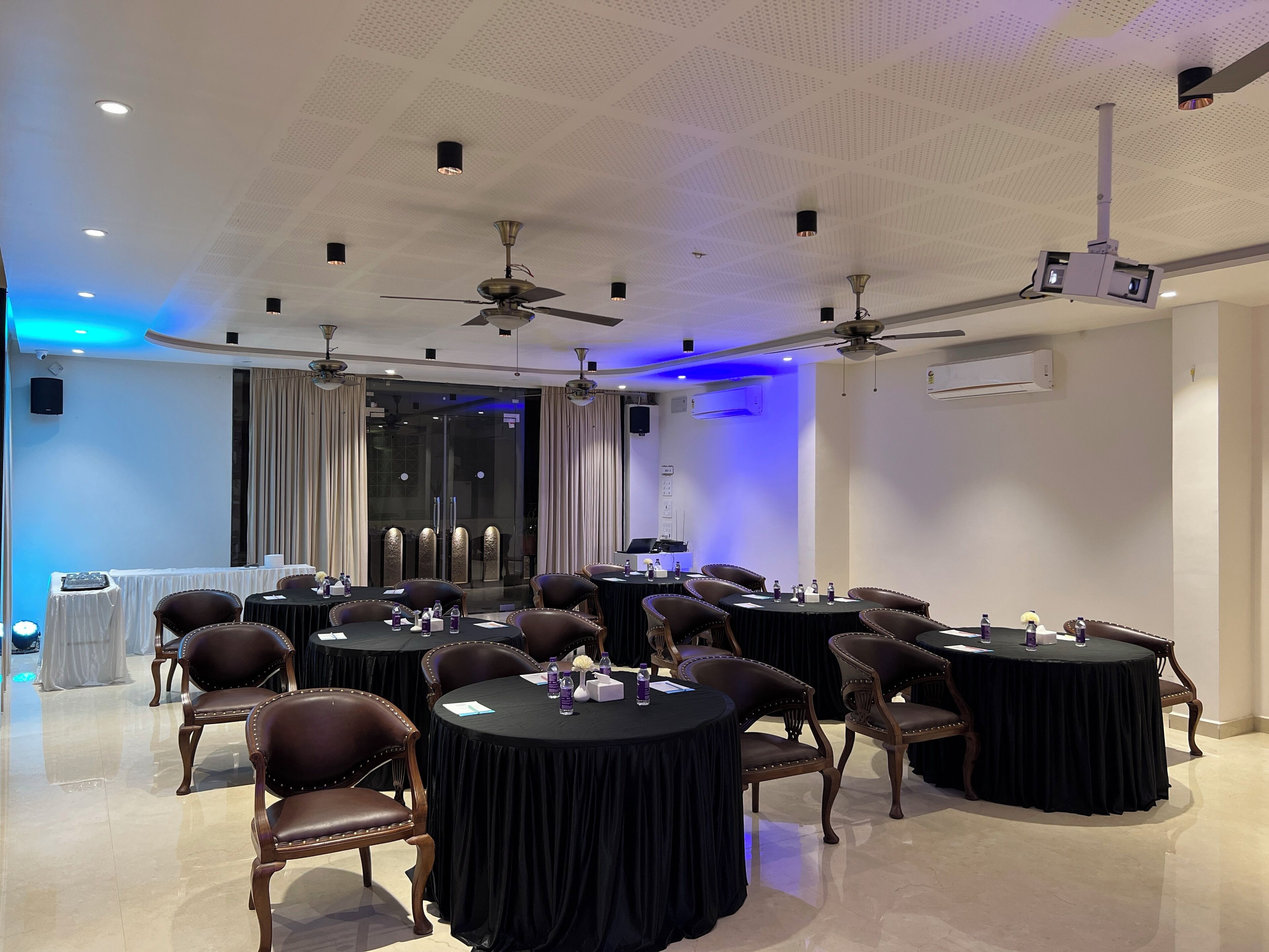 A conference room with round tables, chairs, ceiling fans, and a projector.