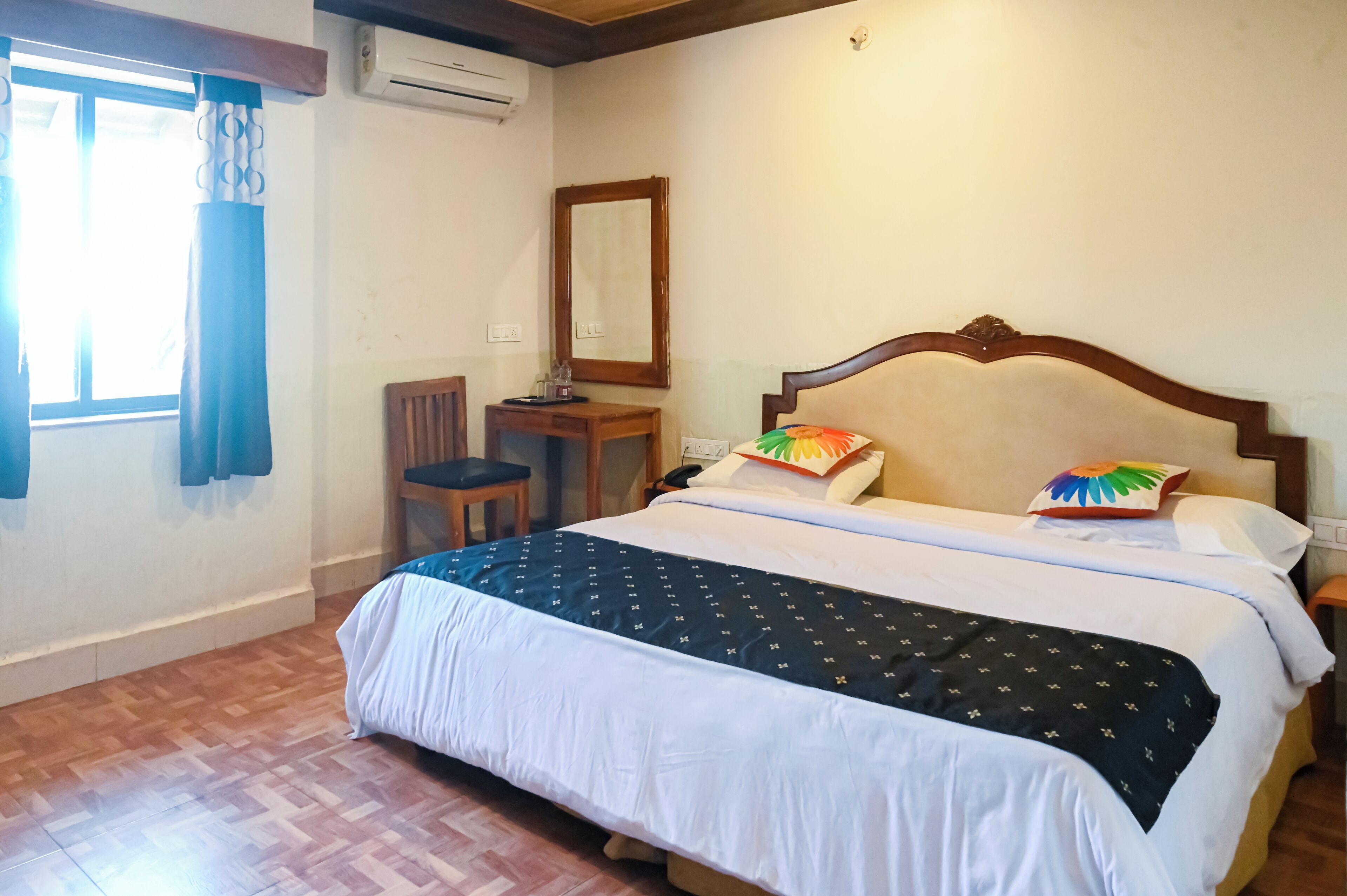 A bedroom with a large bed, a wooden headboard, a bedside table, a chair, a mirror, and a window with curtains.
