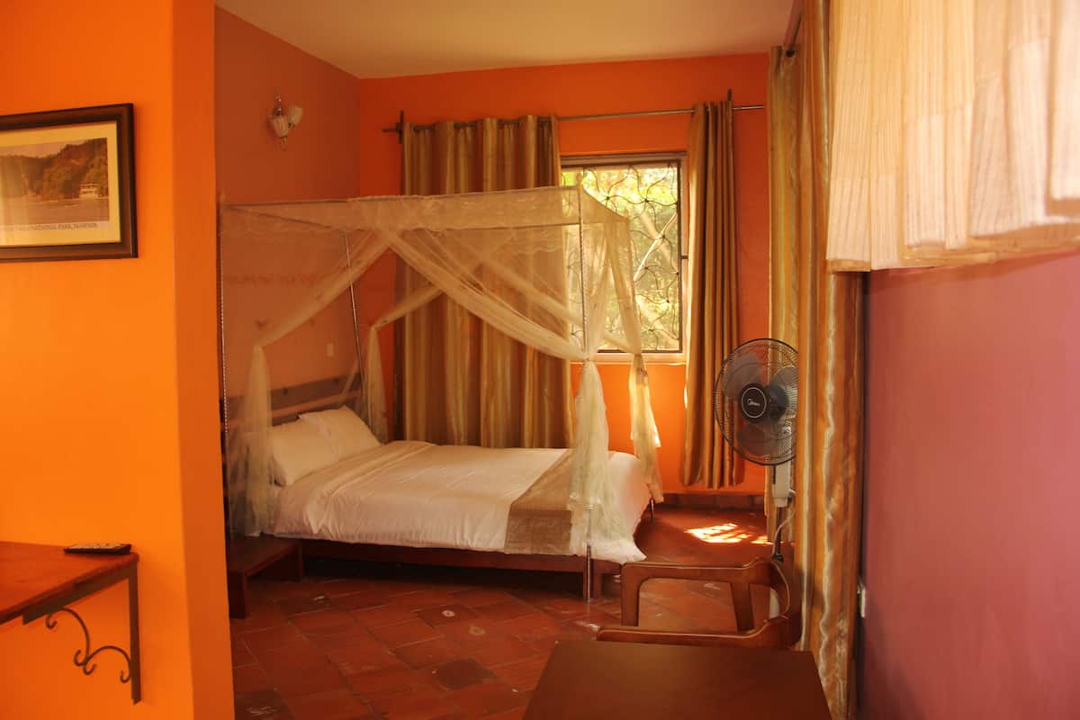 A room with a bed under a mosquito net, a bedside table, a fan, and a window with curtains.