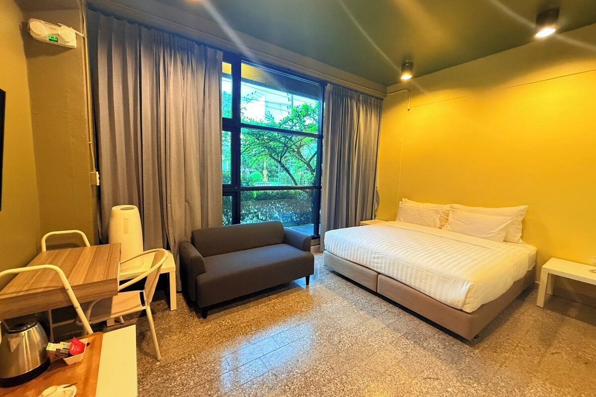 A hotel room with a bed, a sofa, a desk with a kettle, and a window with curtains.