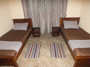 Une chambre double avec deux lits simples, une petite table de chevet en bois, un tapis à rayures et des rideaux à motifs.