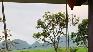 A wooden table with an open book and a pen, a swing chair, and a view of green fields and mountains. - Hung Phat Bungalow (Phong Nha)