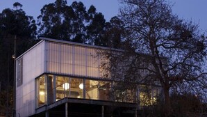 A modern, elevated building with large windows and a flat roof, surrounded by trees.