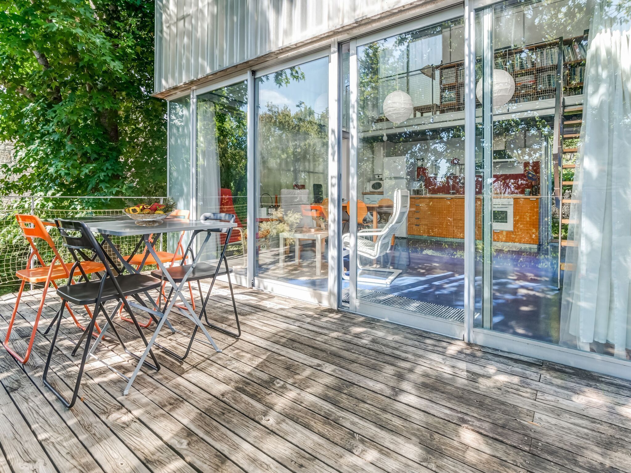 A modern outdoor deck with a glass enclosure, featuring a dining area with orange chairs and a table, and a view into the interior of the building.