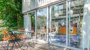 A modern outdoor deck with a glass enclosure, featuring a dining area with orange chairs and a table, and a view into the interior of the building.