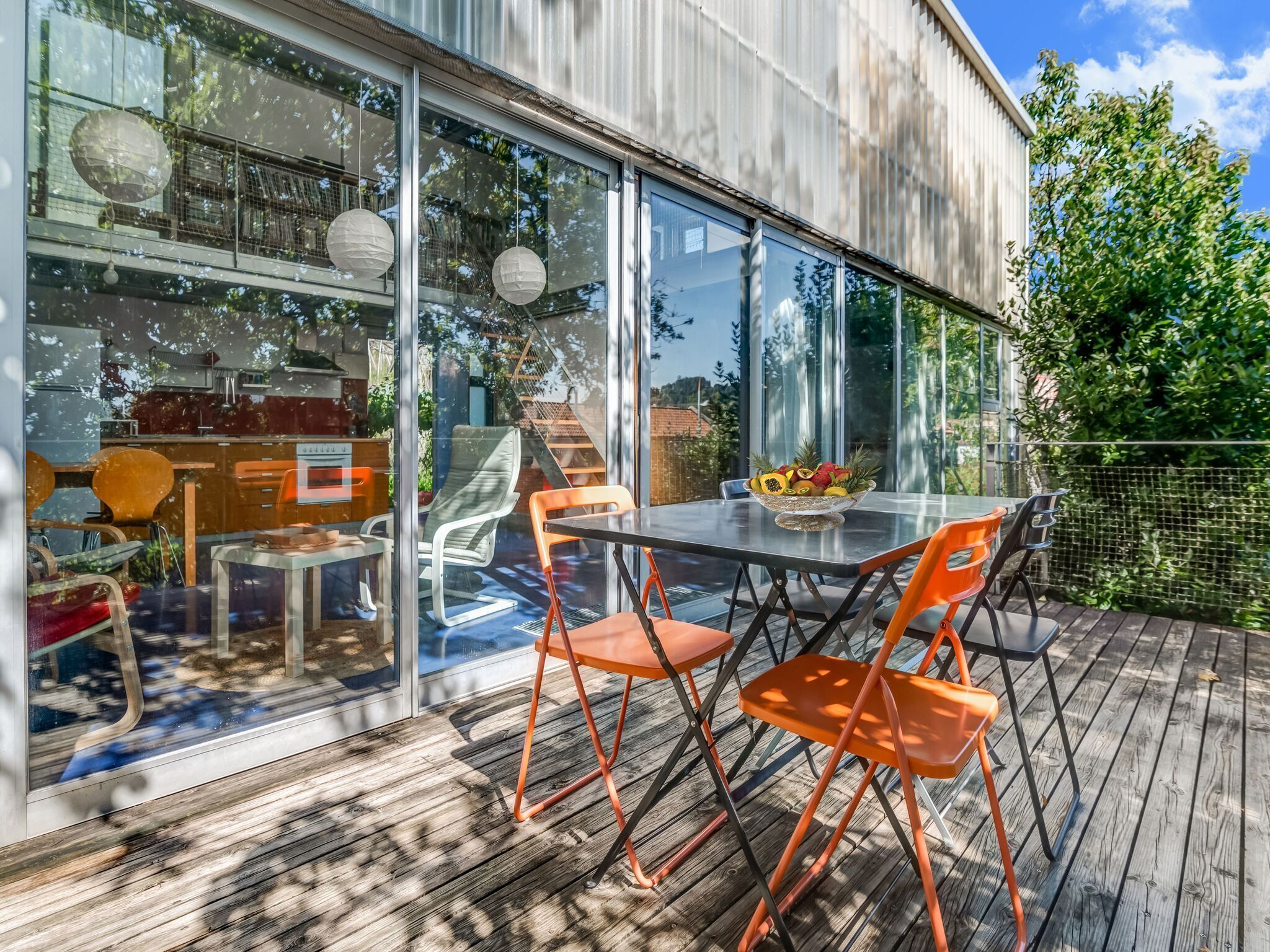 A modern outdoor dining area with a glass wall, wooden deck, and orange chairs.