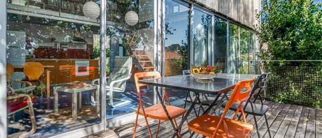 A modern outdoor dining area with a glass wall, wooden deck, and orange chairs.