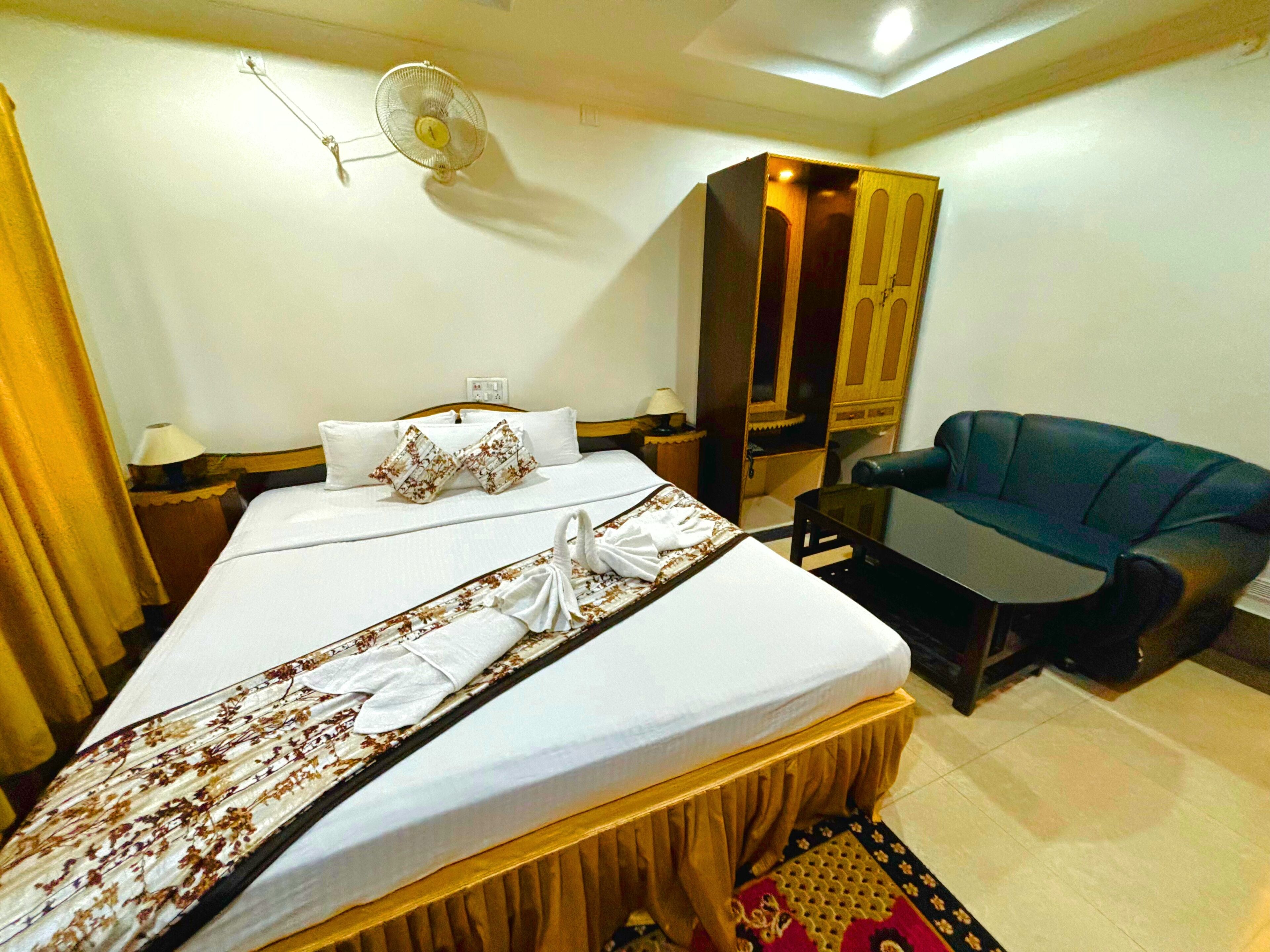A hotel room with a bed, a desk, a chair, a wardrobe, and a wall-mounted fan.