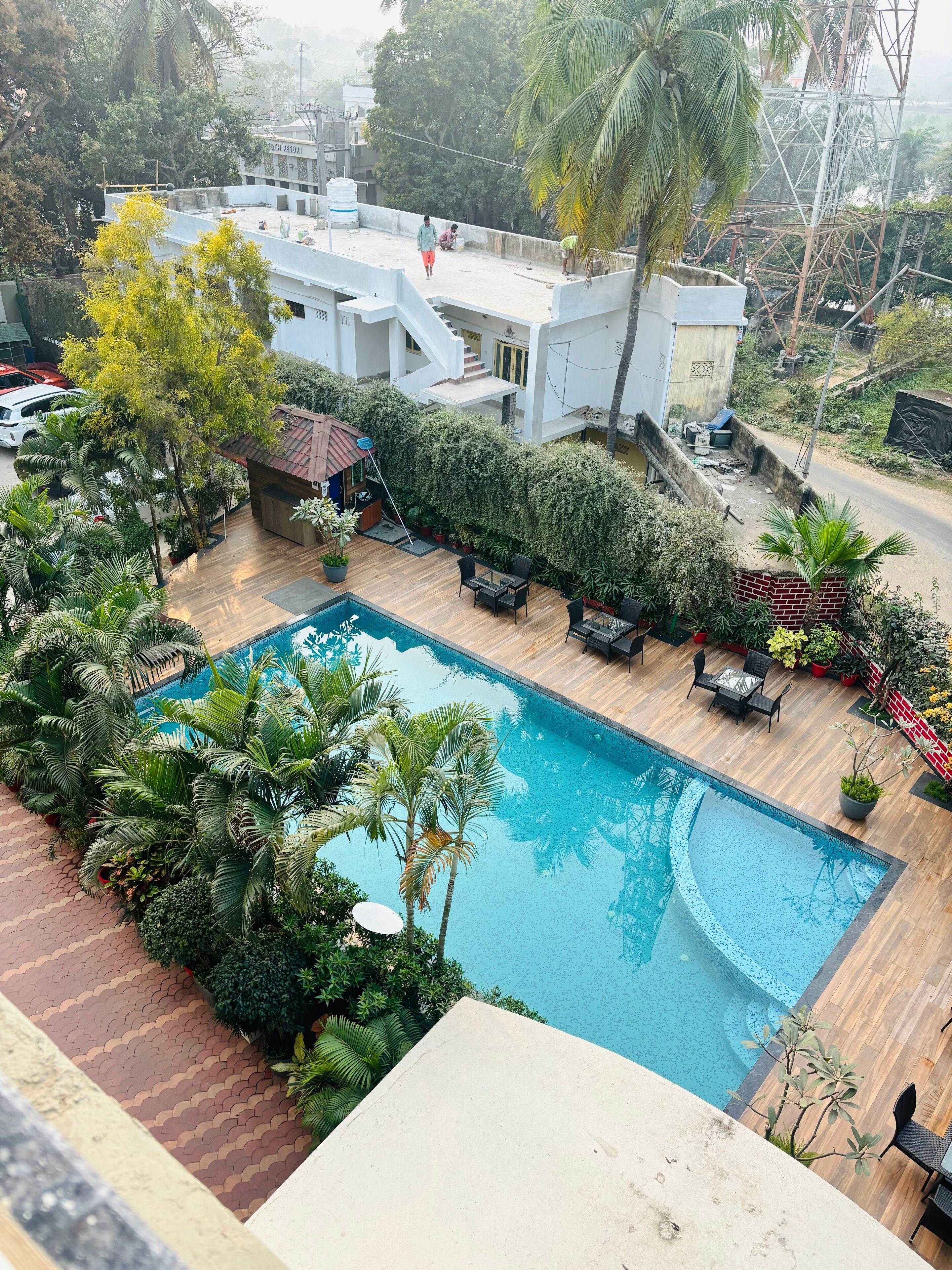 A rooftop pool with wooden deck seating, surrounded by lush greenery and palm trees.