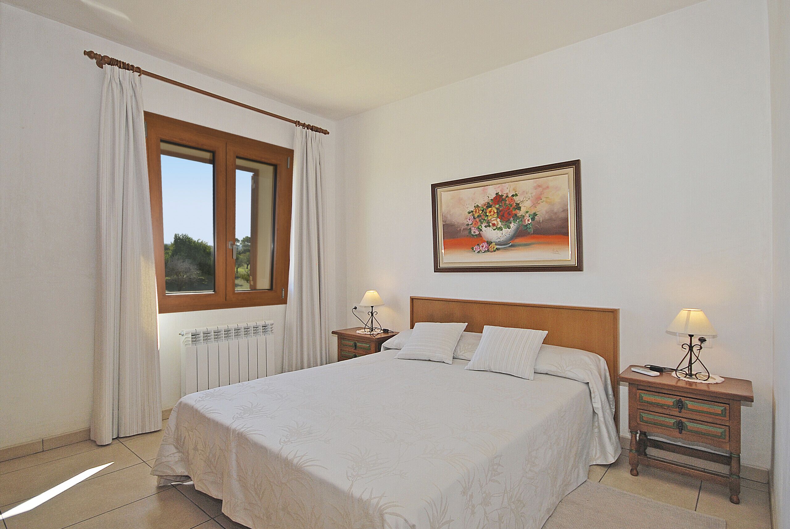 A bedroom with a large bed, two bedside tables, a window with curtains, a radiator, and a painting on the wall.