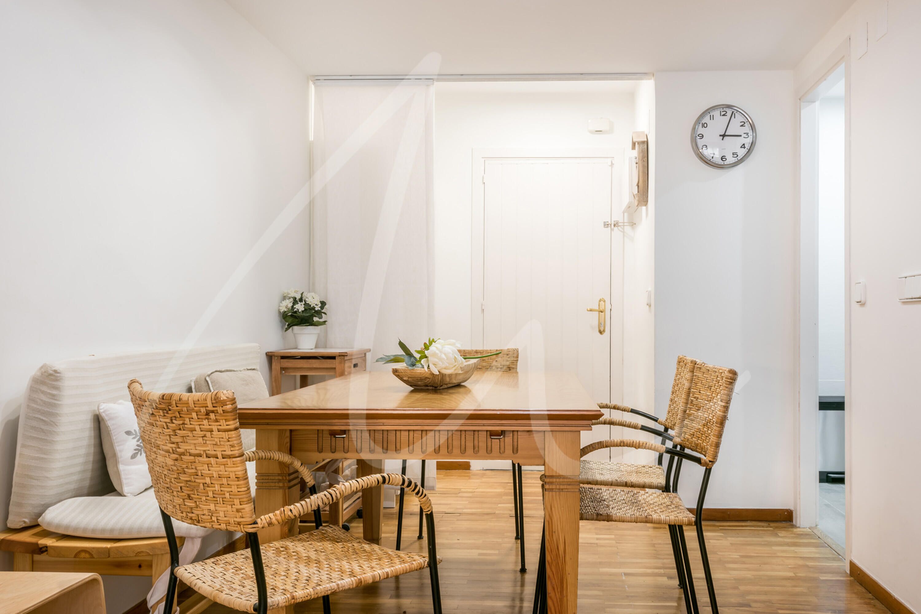 Un comedor con una mesa de madera, sillas de mimbre, un reloj blanco en la pared y una puerta al fondo.