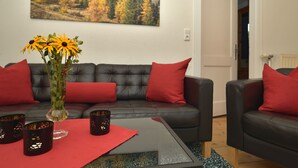 A living room with a black leather sofa, red cushions, a glass coffee table, and a vase with yellow flowers. - Cozy cottage with large garden, balcony, veranda  (Reutte)