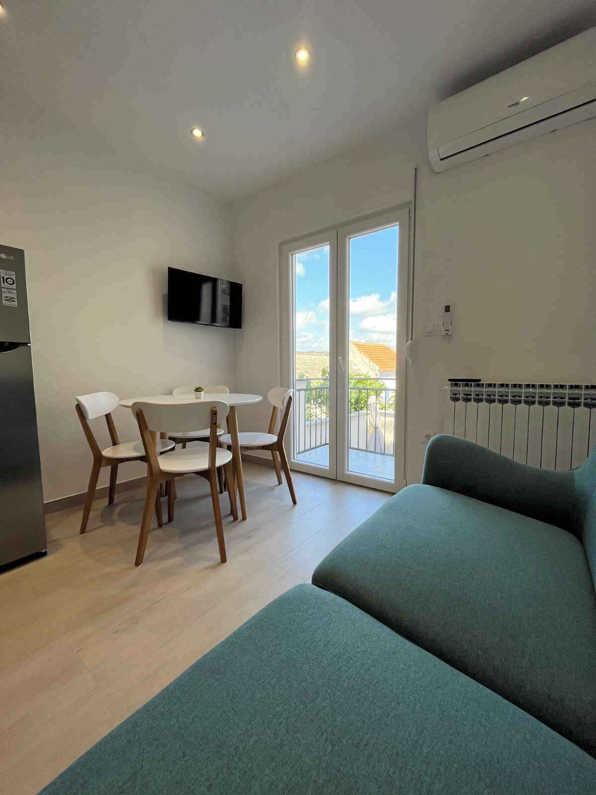 Un salon moderne comprenant un canapé, une table à manger avec des chaises, un téléviseur à écran plat fixé au mur et une grande fenêtre donnant sur l’extérieur.