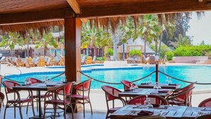 Un restaurante junto a la piscina con mesas al aire libre, piscina y jardines tropicales.