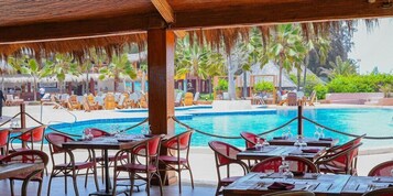Un restaurante junto a la piscina con mesas al aire libre, piscina y jardines tropicales.