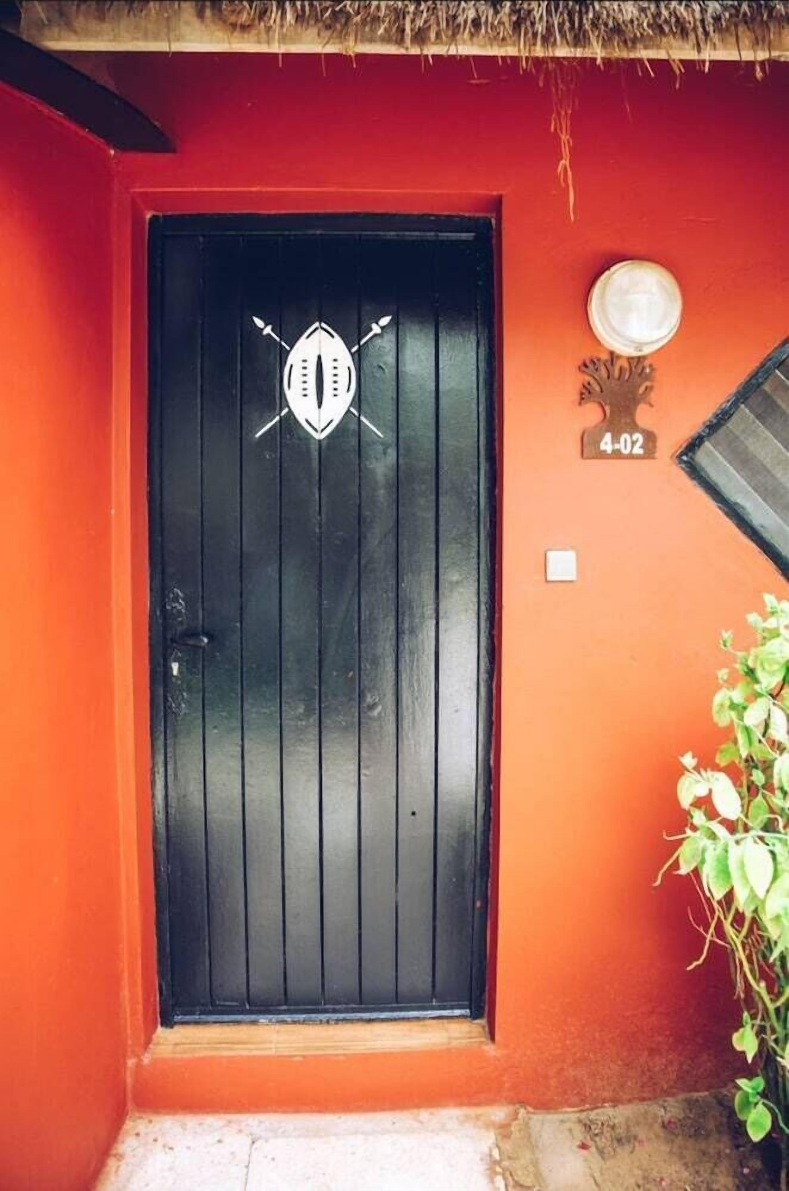 Una puerta negra con un emblema blanco, una pequeña placa con el número "4-02" y una planta en maceta a la derecha.