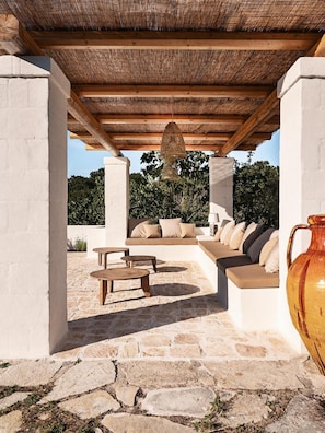 Un patio al aire libre con suelo de piedra, una pérgola de madera, una zona de estar con cojines y un jarrón grande.
