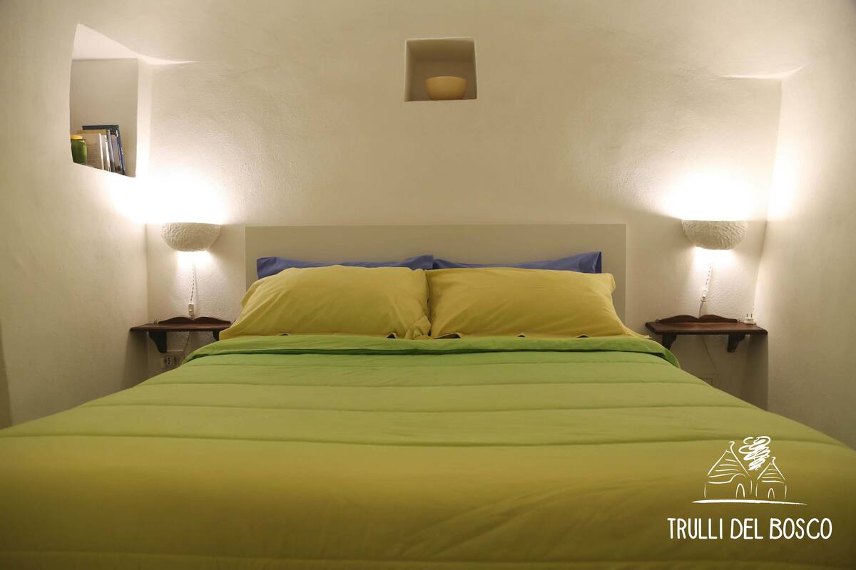 Una cama con una colcha verde y amarilla, dos lámparas de noche y un pequeño estante con libros.