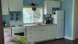 A compact kitchen with white cabinets, a stove, and a refrigerator. There is a window with blinds, a ceiling fan, and a couch with green pillows. - Ocean front cottage in the heart of Hubbards. (Hubbards)