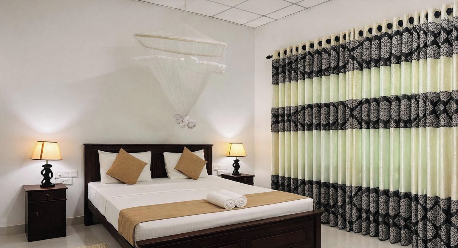 Een hotelkamer met een bed, twee nachtkastjes met lampen, een hangend klamboes en kamergordijnen met een patroon.